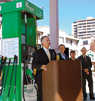 Photo of four men in suits standing behind a podium , giving a press conference at a fueling station in front of a fuel pump that dispenses ethanol and biodiesel.