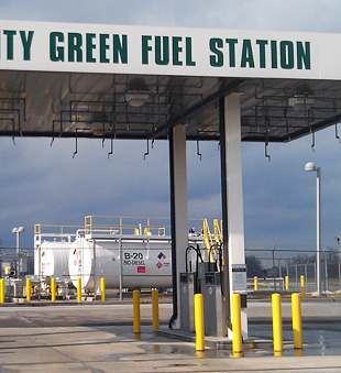 Photo of a fueling station with large biodiesel tanks in the background. A sign on the fuel canopy reads, Monroe County Green Fuel Station.