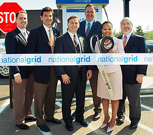 Photo of a group of men and women standing in front of a compressed natural gas (CNG) fueling station for a ribbon-cutting ceremony.