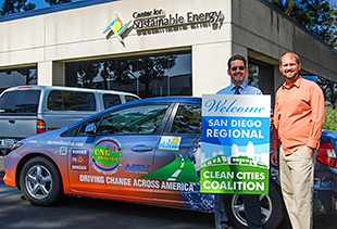 A photo of two men posing in front of a vehicle wrapped in graphics that market Clean Cities, holding a sign that says, "Welcome San Diego Regional Clean Cities Coalition."