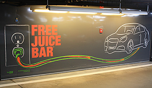 Photo of black wall in a parking garage with a mural of a car plugged into an outlet that says, "Free Juice Bar."