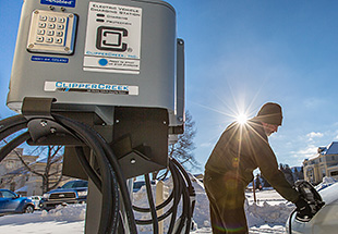 A photo of a man charging an electric vehicle in the snow with the sun shining in the background.