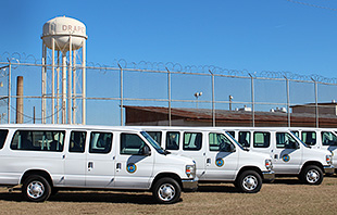 Picture of five white propane-fueled vans in front of prison with barbed wire fence and water tower that says "Draper." 